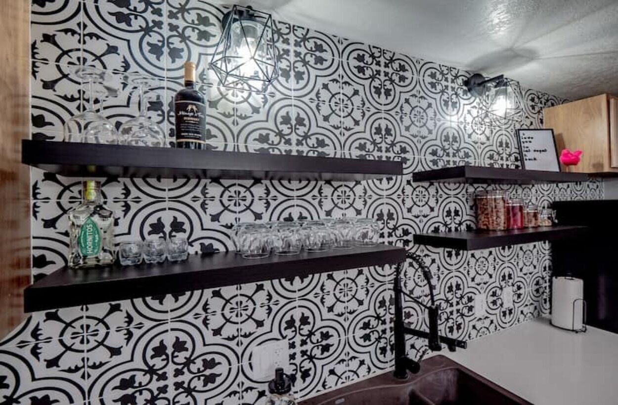 Uma cozinha com papel de parede estampado, prateleiras com copos e garrafas, e um lustre.