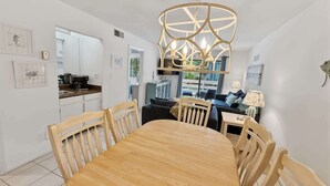 A dining area with a wooden table and chairs, a hanging light fixture, and a kitchen in the background. - Renovated Beach Villa 14F: Gated Community near Golf and Bike Trails (Hilton Head Island)