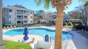A pool area with lounge chairs, a palm tree, and a building with balconies. - Renovated Beach Villa 14F: Gated Community near Golf and Bike Trails (Hilton Head Island)