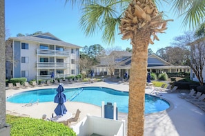 A pool area with lounge chairs, a palm tree, and a building with balconies. - Renovated Beach Villa 14F: Gated Community near Golf and Bike Trails (Hilton Head Island)
