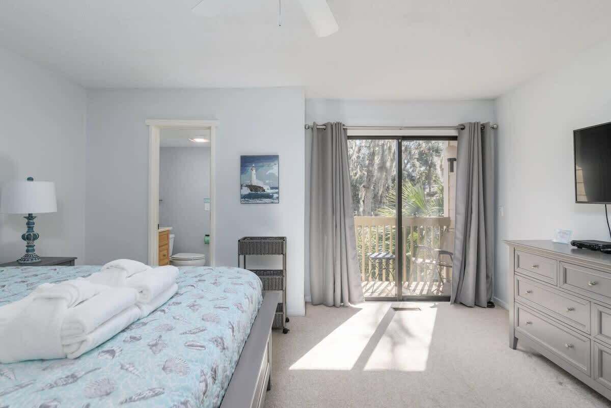 Een slaapkamer met een bed, een dressoir, een televisie, een schilderij en een balkon met gordijnen.
