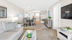 A living room with a sofa, coffee table, TV, dining area with chairs, and a kitchen in the background. - Amazing Ocean Views From Your Newly Renovated Oceanfront Condo in Carolina Beach (Carolina Beach)
