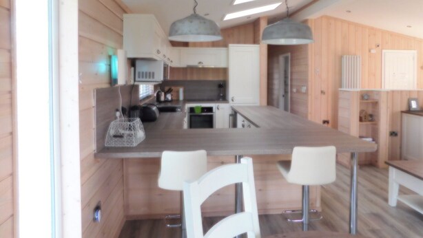 A modern kitchen with wooden walls, a central island with bar stools, pendant lighting, and built-in appliances. - Spacious 2-bed Lodge in Bury Saint Edmunds (Bury St Edmunds)