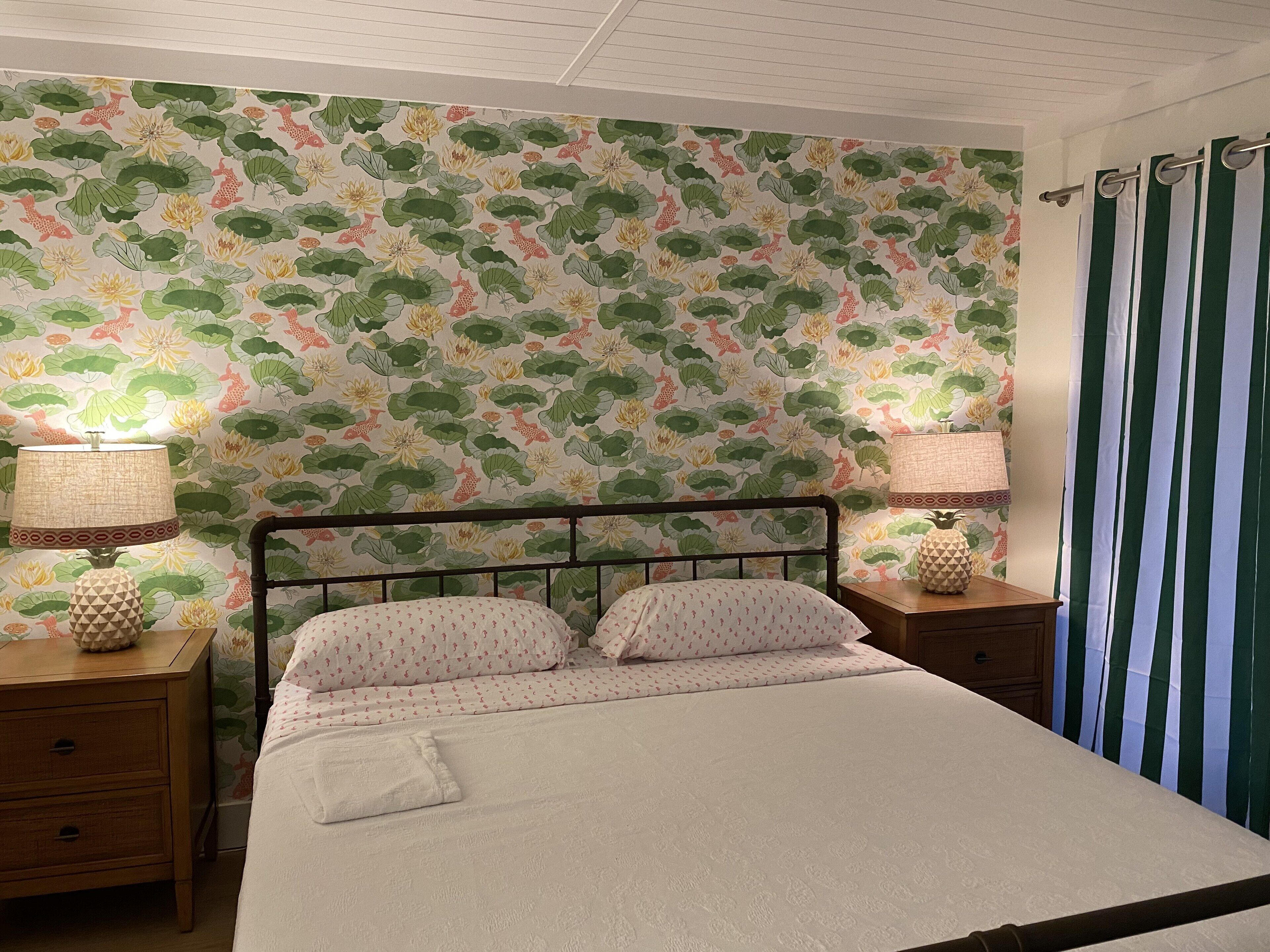 A bedroom with a floral wallpaper, a bed with white bedding, two bedside tables with lamps, and a striped curtain.