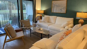Moderni olohuone, jossa on valkoinen sohva, rottinkituoli ja lasinen sohvapöytä. Ikkunasta avautuu näkymä vihreälle seinälle, ja sohvan yläpuolella on taulu.