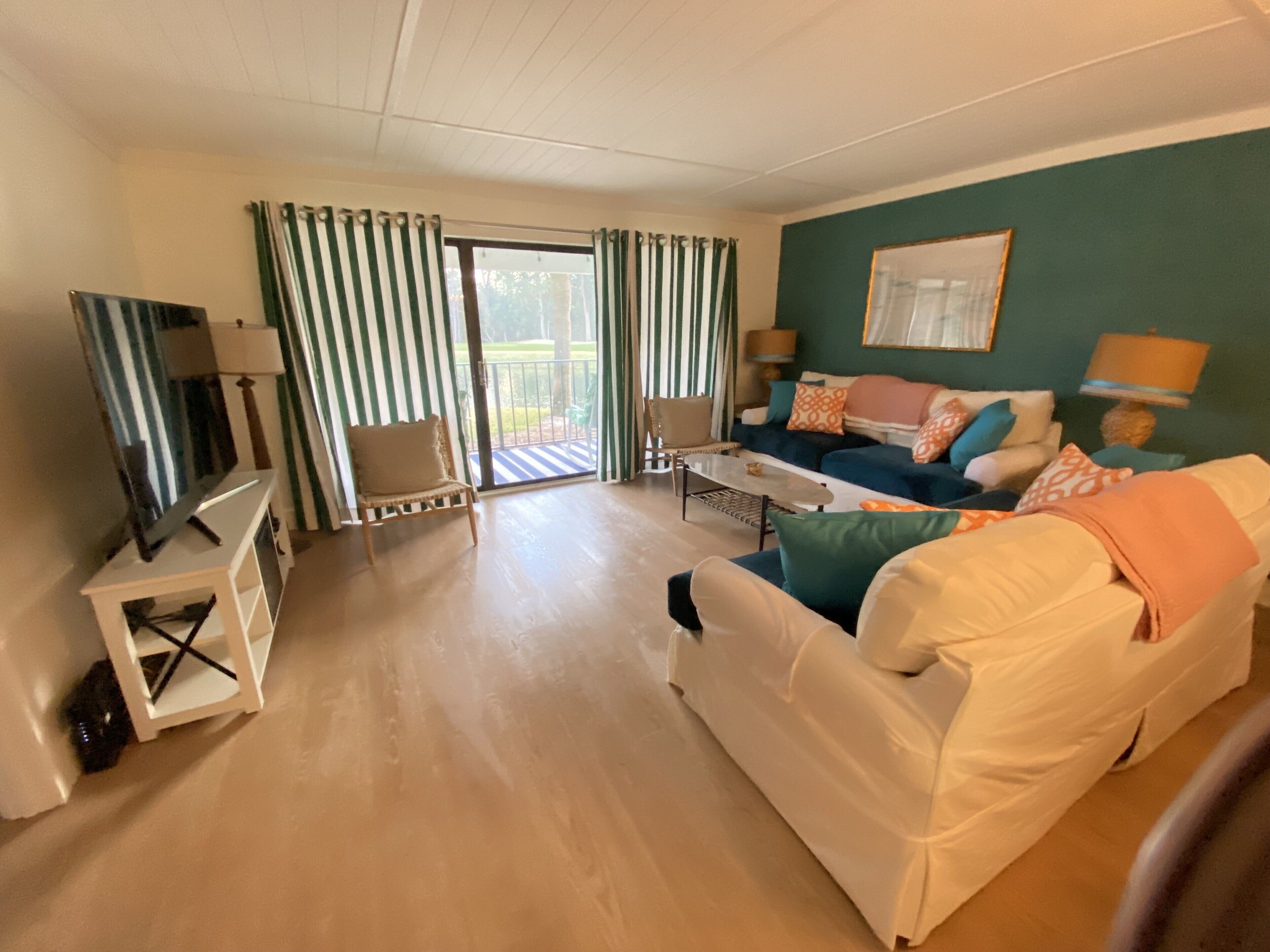 Sala de estar con sofá, televisión, mesa de centro y una silla. Hay un ventanal corredero que da a un espacio exterior.