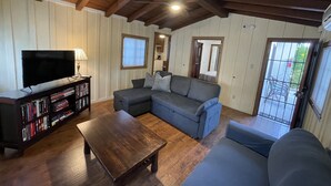 A living room with a sectional sofa, a coffee table, a TV on a stand, and a bookshelf. - Villa M - 1bedroom villa with kitchen, steps from downtown Palm Springs! 🍒 (Palm Springs)