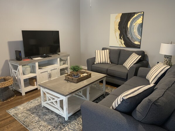 A living room with a grey sofa, a white coffee table, a TV on a white stand, and a painting on the wall.