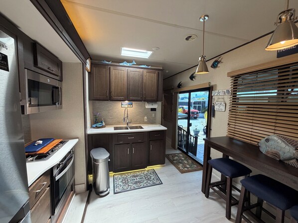 A compact kitchen with dark wooden cabinets, stainless steel appliances, and a small dining area with bar stools. - Calusa Campground #50- RV in Key Largo Florida Keys Vacation Paradise Place (Key Largo)