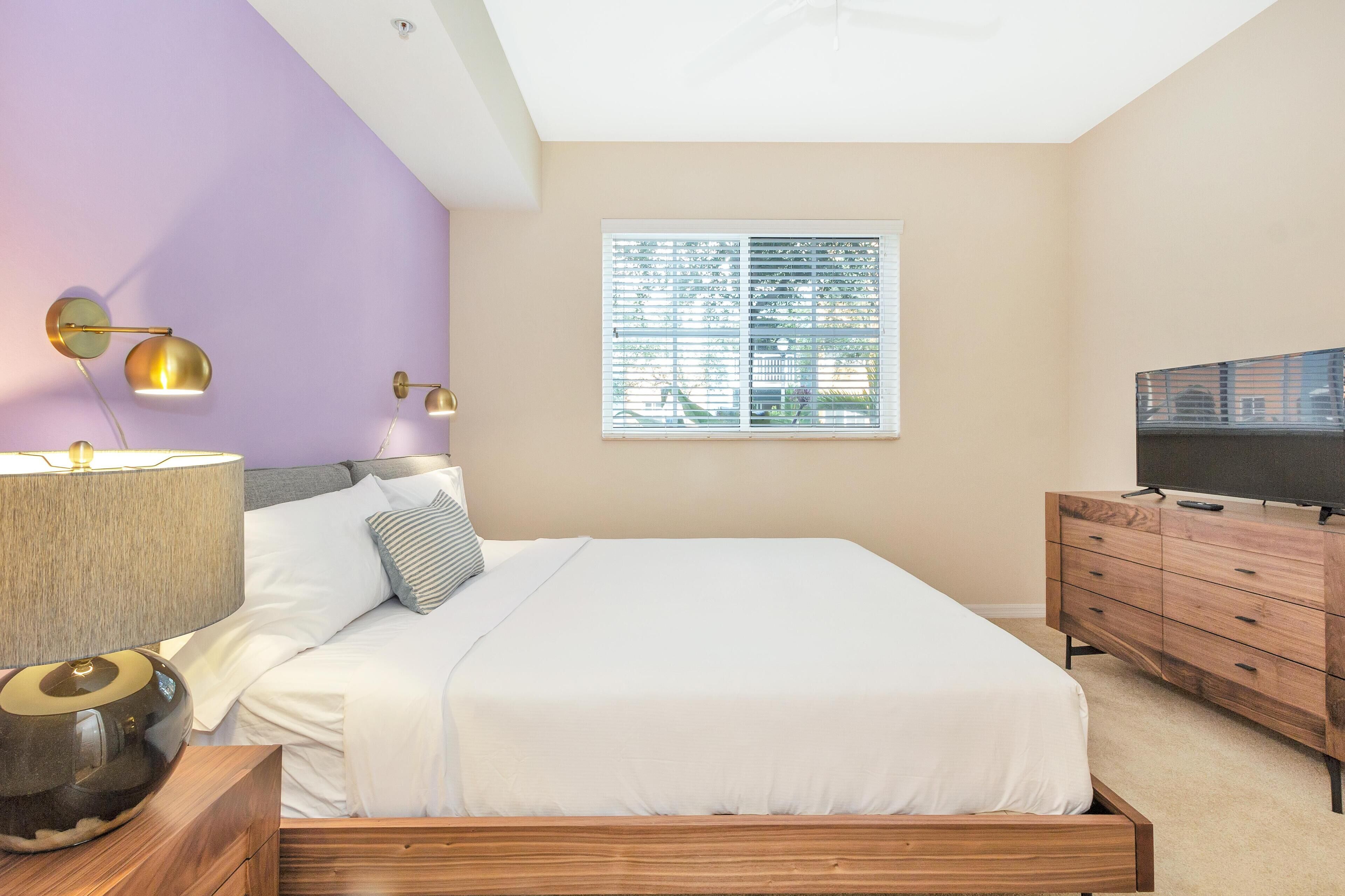 A bedroom with a bed, bedside lamps, a TV on a wooden dresser, and a window with blinds.