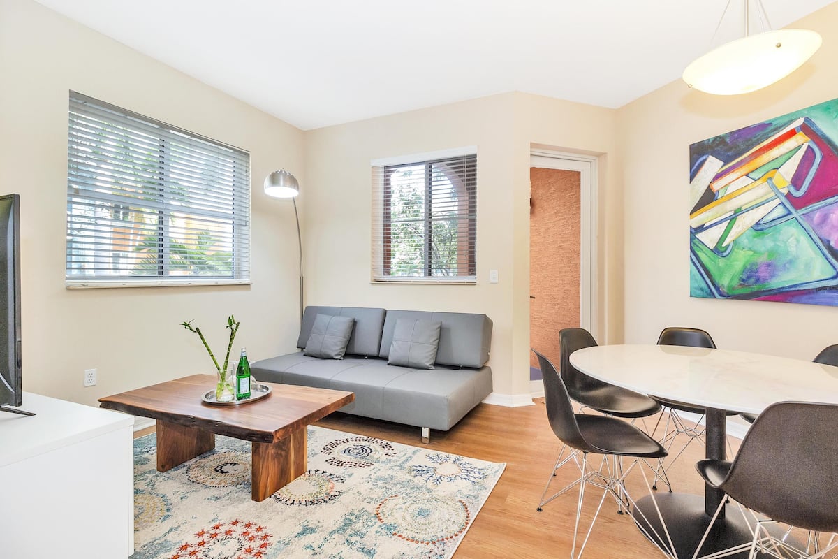A modern living room with a gray sofa, a wooden coffee table, a dining table with chairs, and a colorful abstract painting on the wall.