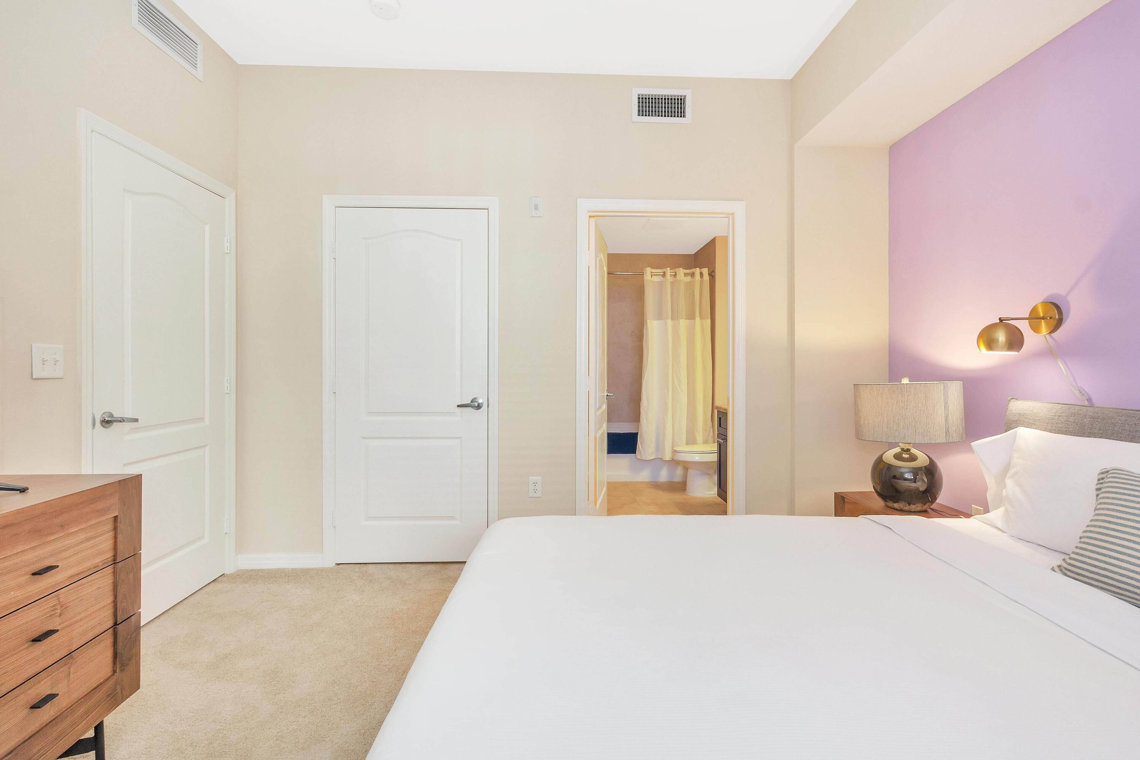 A bedroom with a bed, a dresser, a nightstand with a lamp, and a bathroom visible through an open door.