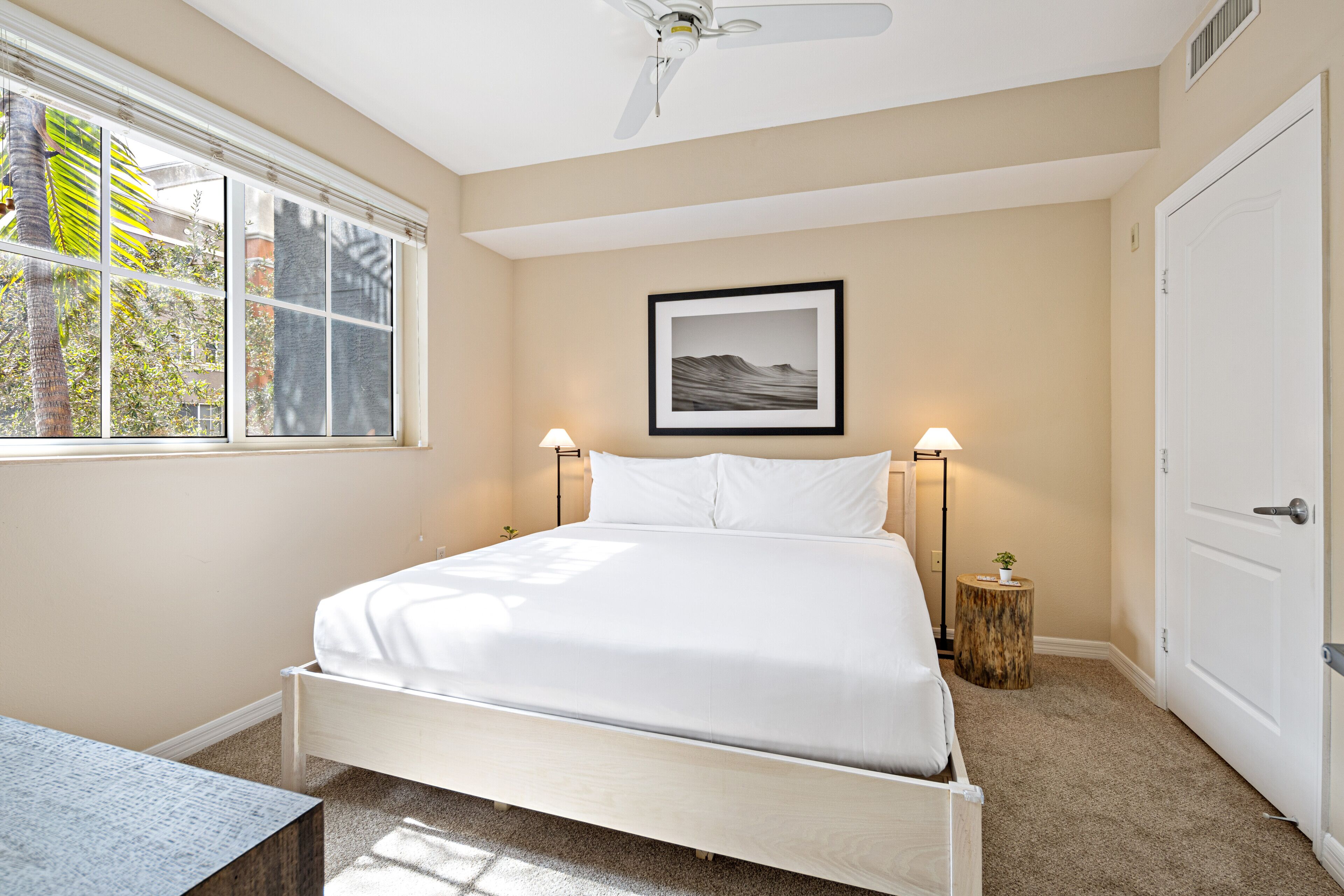 Une chambre à coucher avec un lit, une fenêtre, un ventilateur de plafond, un tableau encadré et une table de chevet avec une lampe.