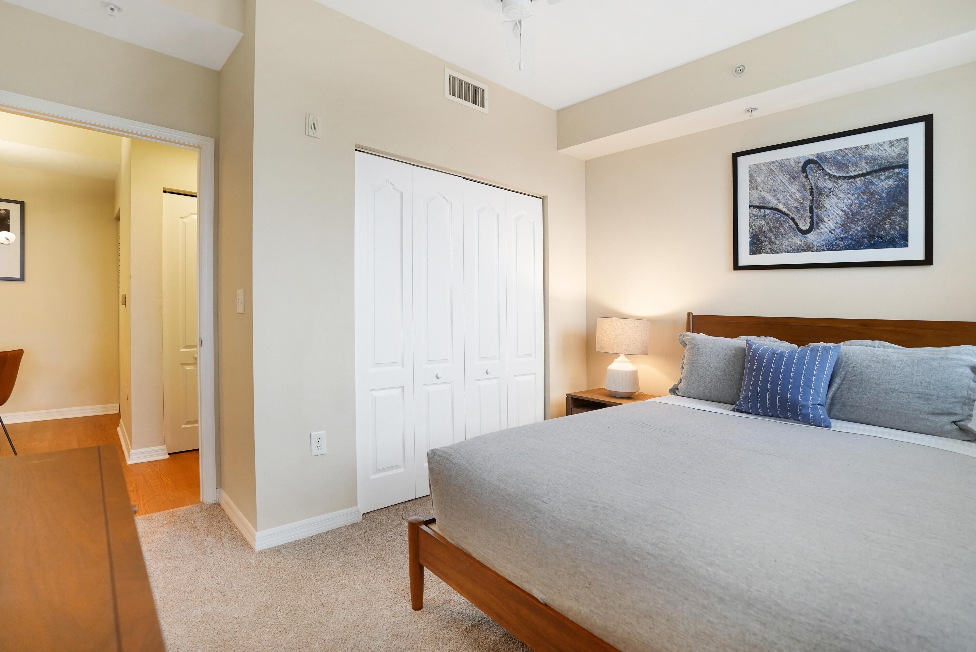 A bedroom with a bed, a nightstand, a lamp, a framed picture on the wall, and a door leading to another room.