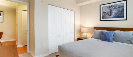 A bedroom with a bed, a nightstand, a lamp, a framed picture on the wall, and a door leading to another room.