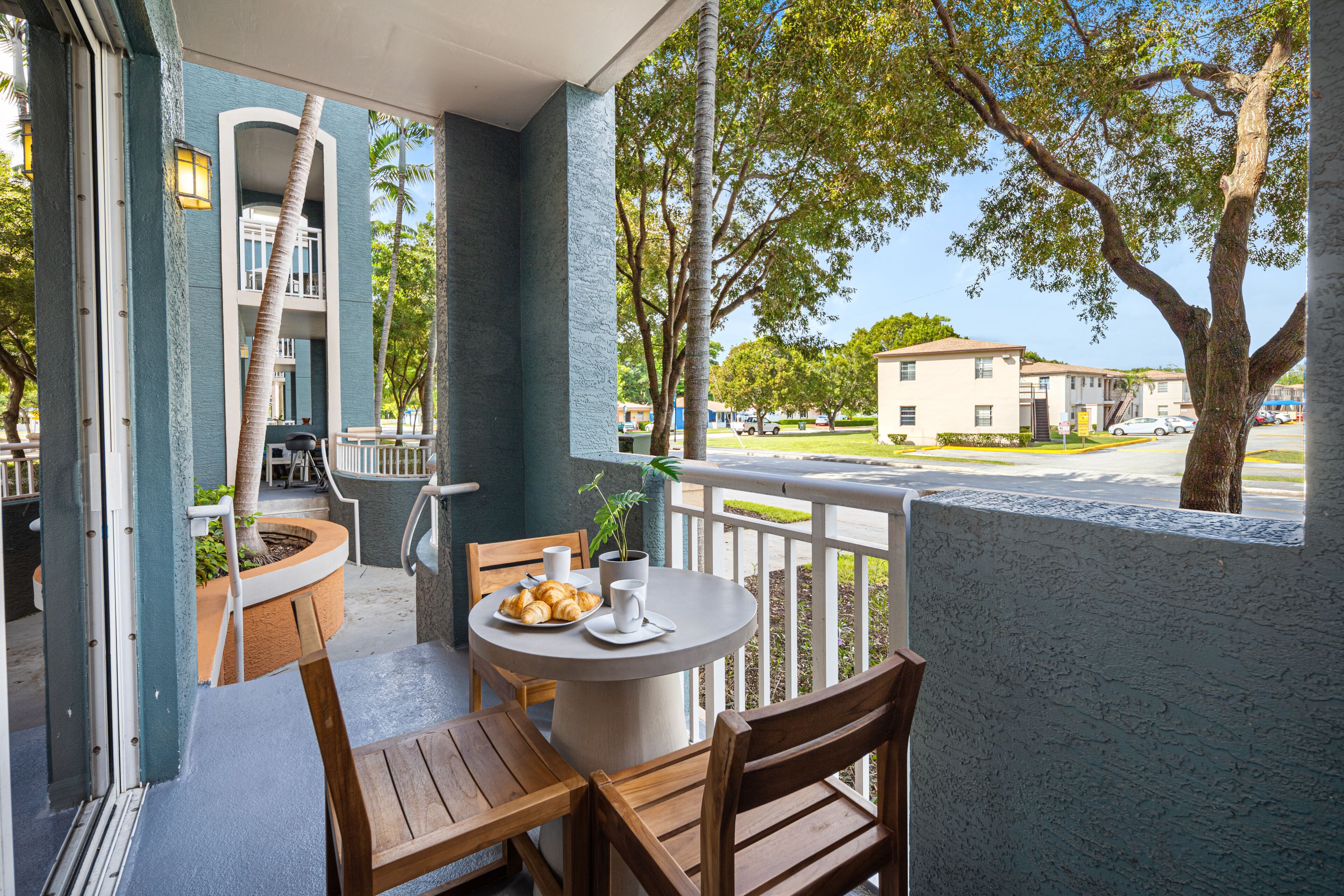 A balcony with a table and chairs, a potted plant, and a view of a residential area with trees and buildings.