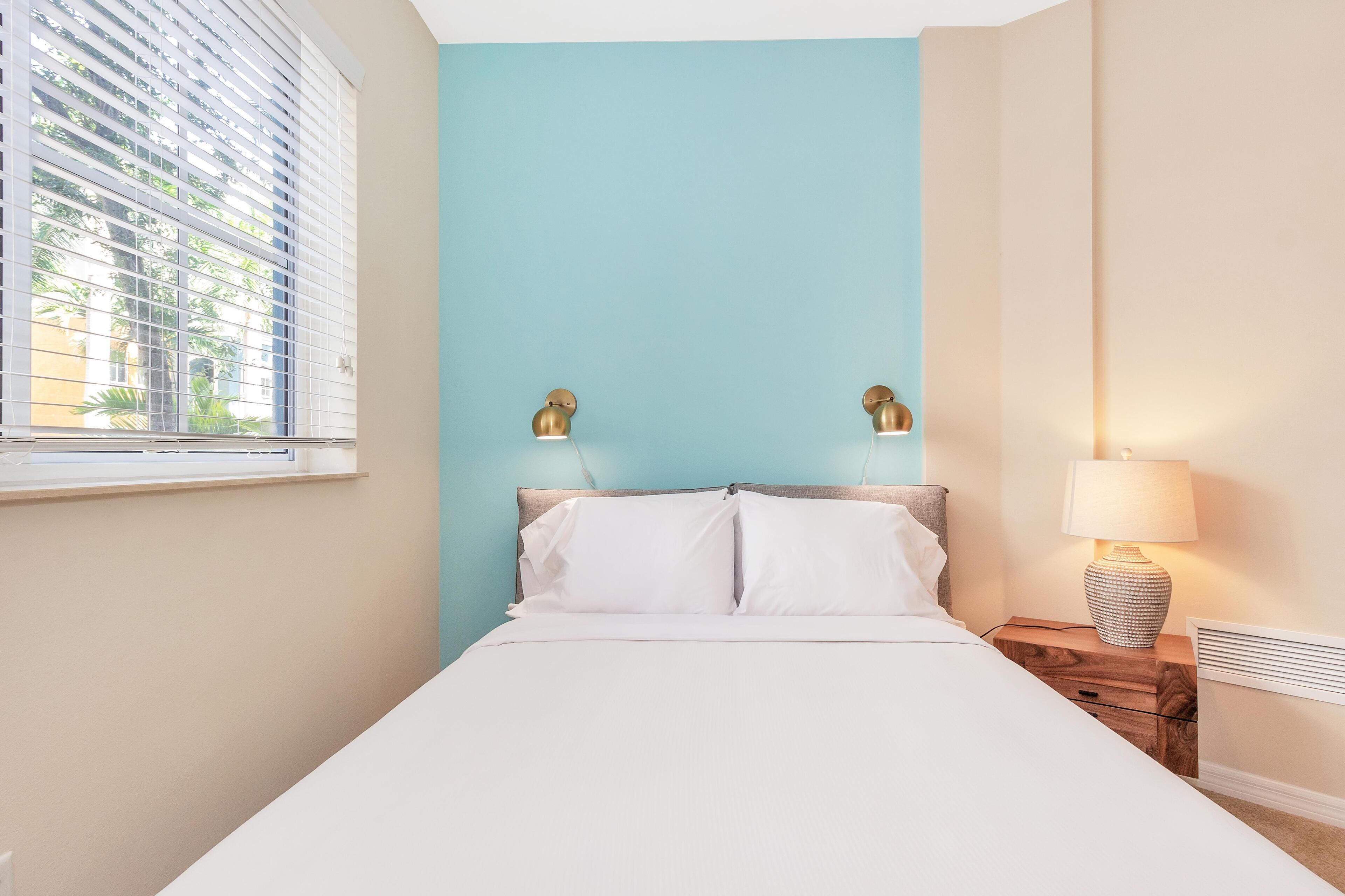 A bedroom with a bed, a nightstand with a lamp, and a window with blinds.