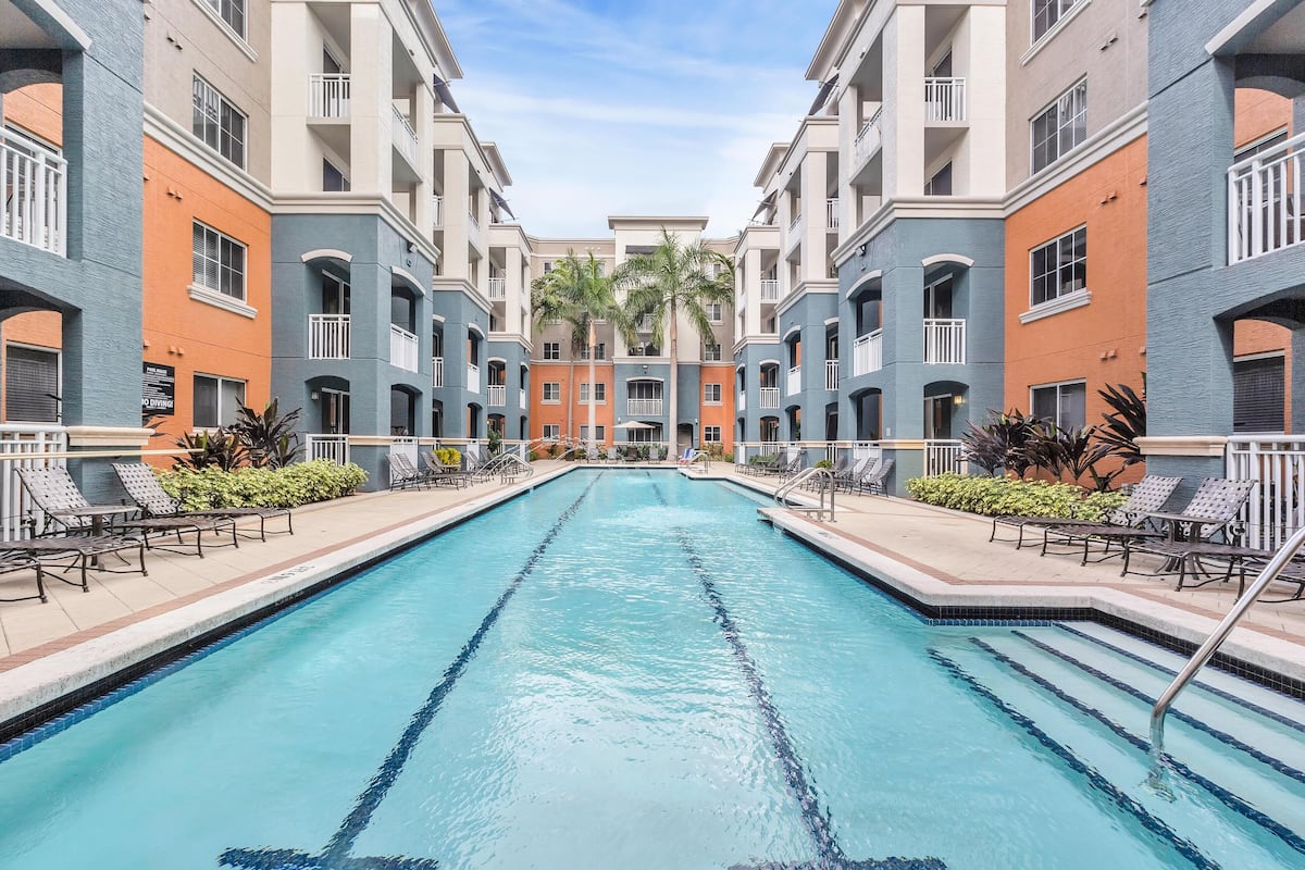 A swimming pool surrounded by multi-story buildings with balconies, palm trees, and lounge chairs.
