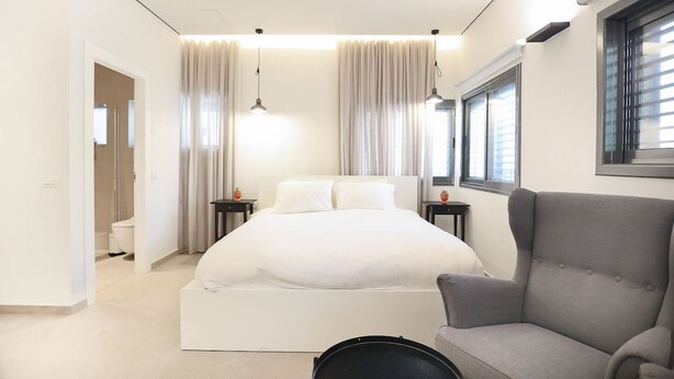 Une chambre moderne avec un grand lit, un fauteuil gris et une fenêtre avec des stores.