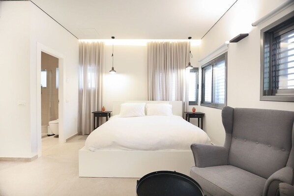 A modern bedroom with a large bed, a grey armchair, and a window with blinds.