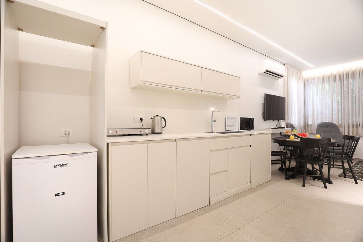 A compact kitchen with a refrigerator, microwave, and kettle, a dining area with chairs and a table, and a TV mounted on the wall.