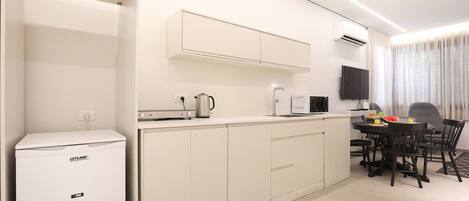 Una cocina compacta con refrigerador, microondas y hervidor, un comedor con sillas y mesa, y un televisor instalado en la pared.