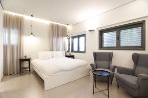 A modern bedroom with a bed, two bedside tables, a chair, a small table, and a window with blinds.
