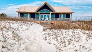 Une maison de plage dotée d’une grande véranda à l’avant, de deux chaises et offrant une vue sur la plage.