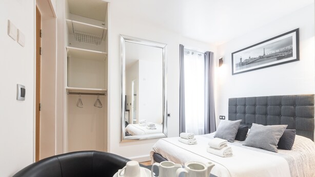 Une chambre d’hôtel avec un lit, une table à manger avec une carafe et un verre, une chaise, une armoire et un tableau encadré au mur.