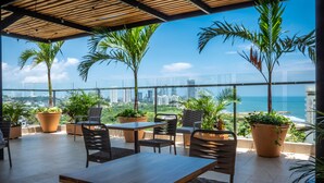 A rooftop terrace with a wooden table and chairs, surrounded by potted plants, overlooking a cityscape and the ocean.