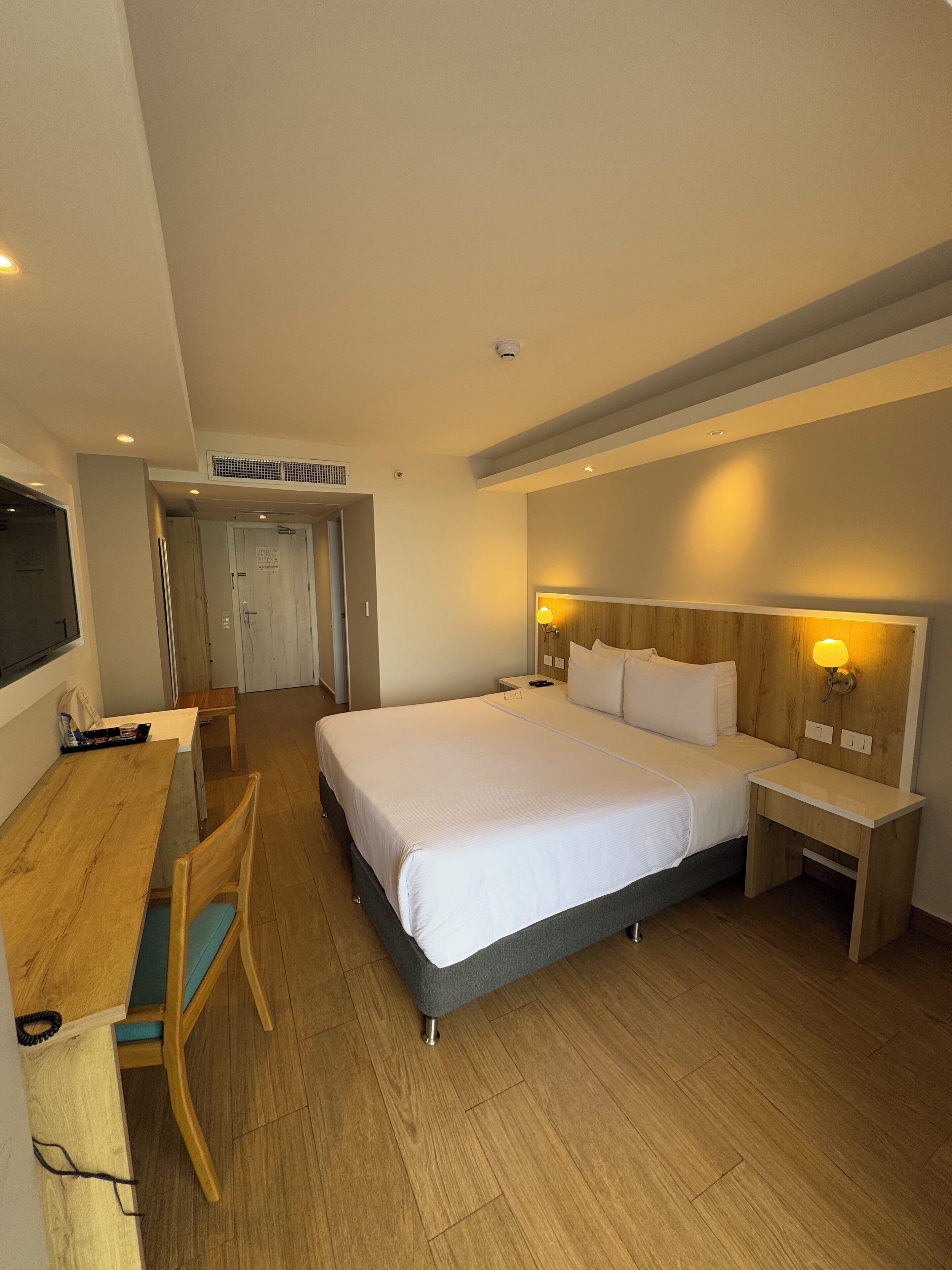 A hotel room with a large bed, bedside tables, a desk with a chair, and a television.