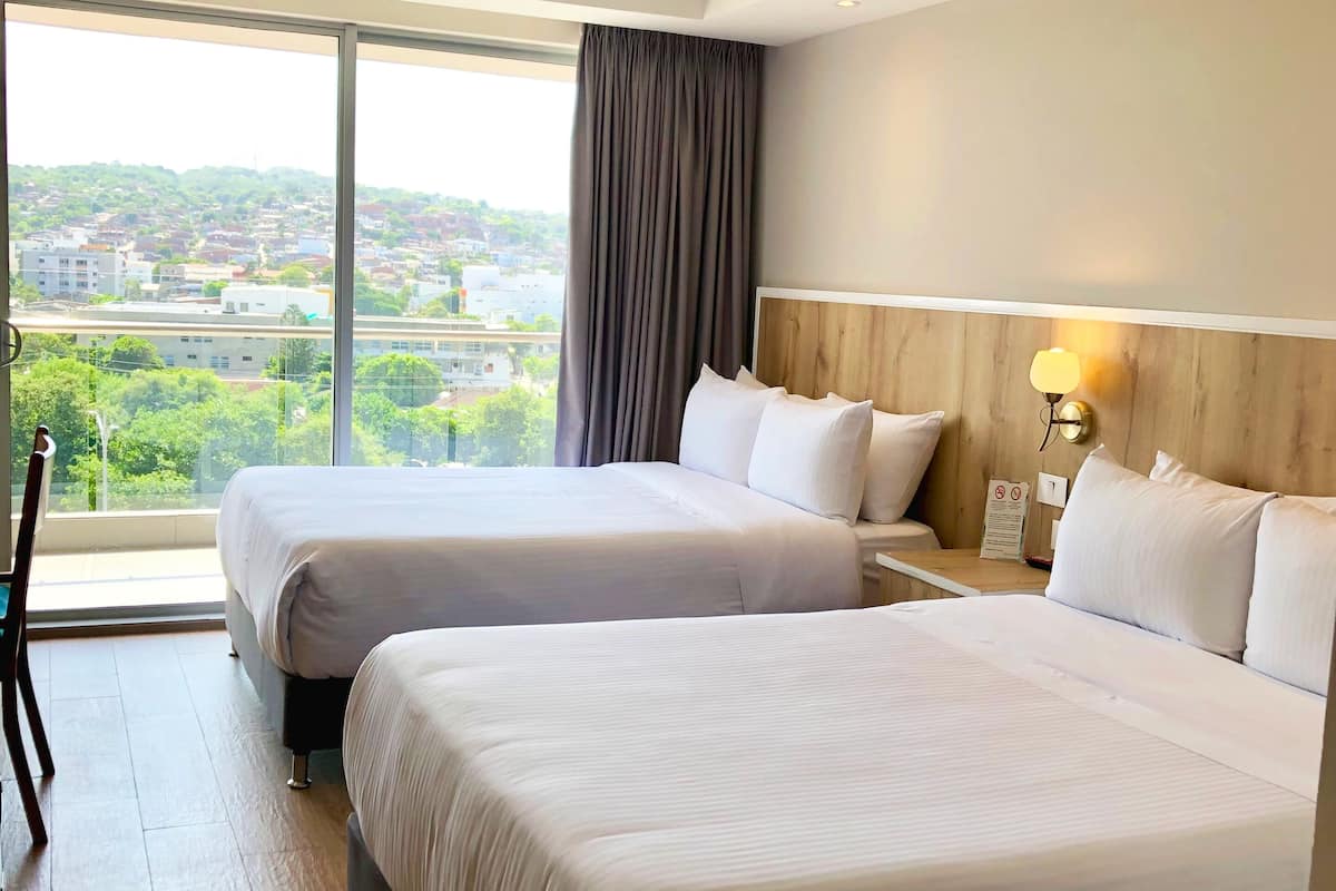 Een hotelkamer met twee bedden, een groot raam met uitzicht op een stadsgezicht, een houten hoofdeinde en een nachtkastje met een lamp.