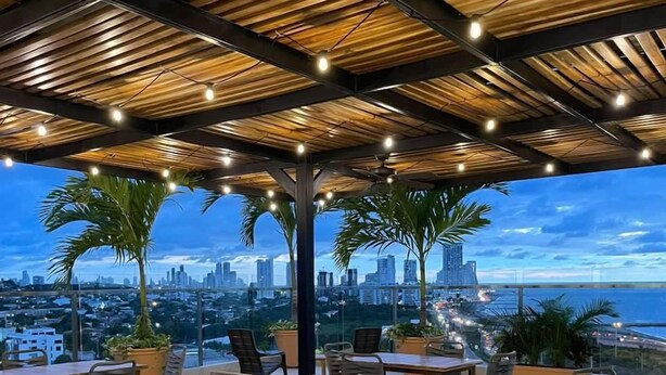 An outdoor dining area with tables and chairs, a city skyline view, and a wooden ceiling structure.
