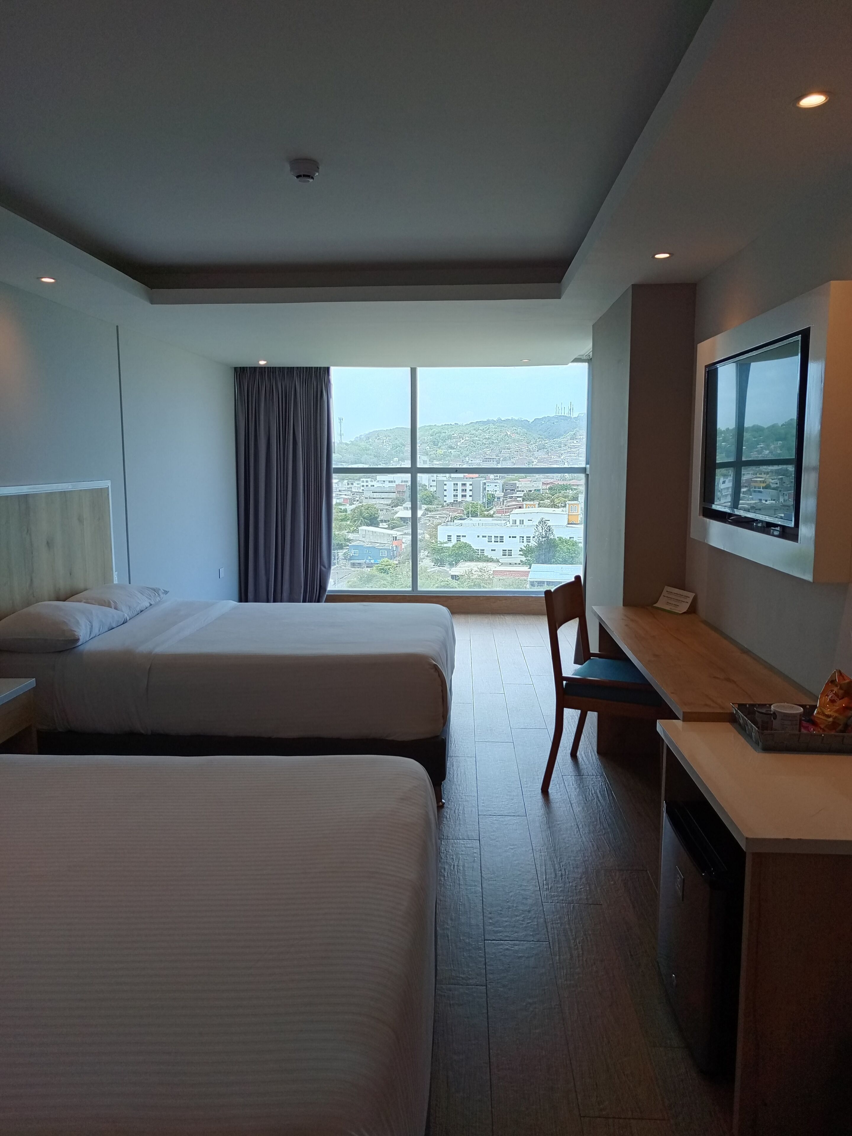 A hotel room with two beds, a desk, a chair, a TV, and a large window offering a city view.