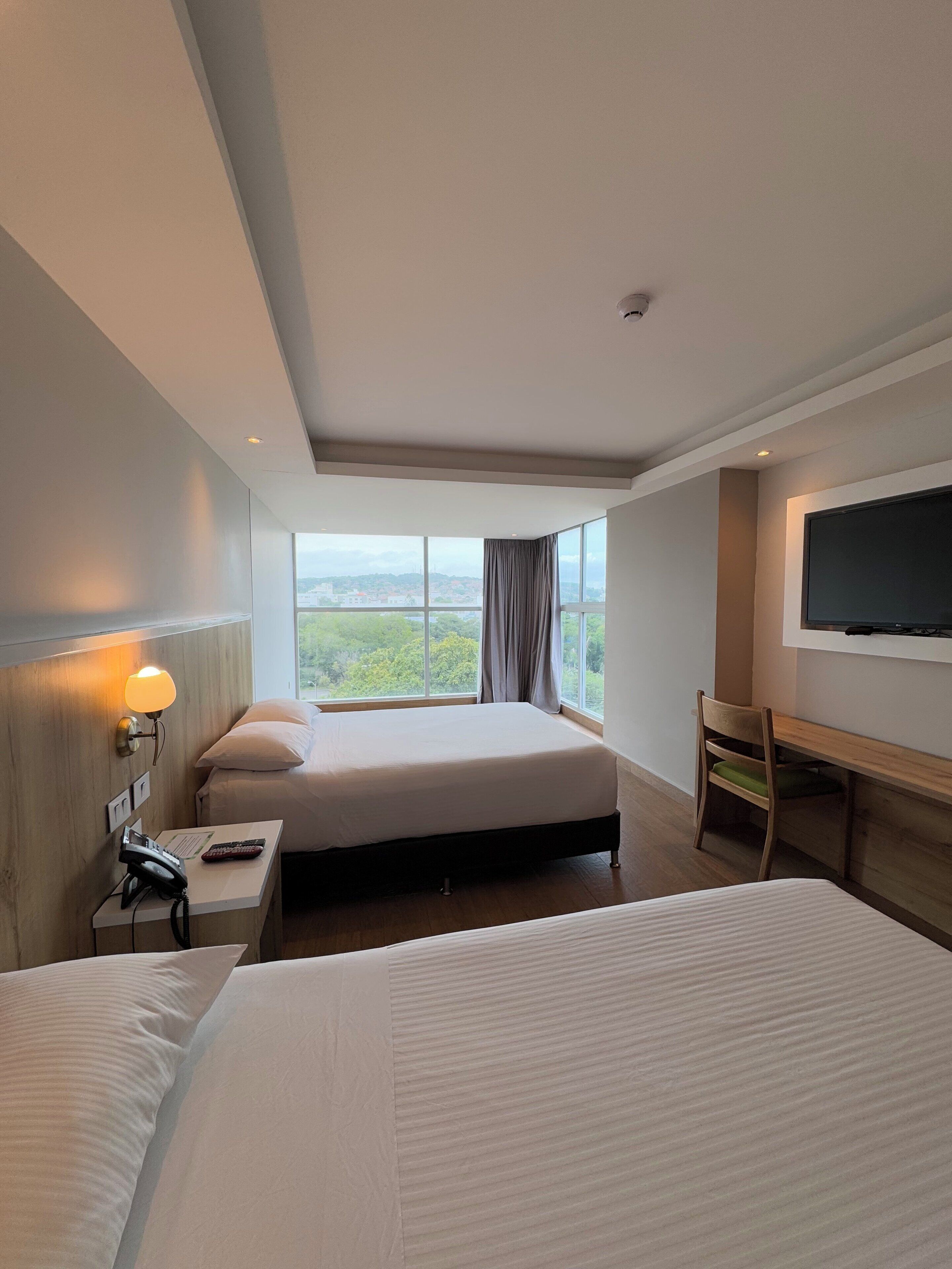 A hotel room with two beds, a desk, a chair, a TV, and a window with a city view.