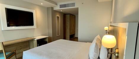A modern hotel room with a large bed, a desk, a TV mounted on the wall, and a bedside lamp.