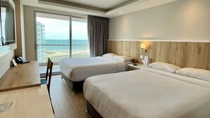A hotel room with two beds, a desk, a TV, and a view of the sea.