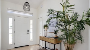 Un pasillo limpio y moderno con una puerta blanca, una consola de madera con un jarrón y una planta, y una planta grande en maceta.
