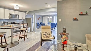 Eine moderne Küche mit weißen Schränken, einer Edelstahl-Mikrowelle und einem Essbereich mit rundem Tisch und Barhocker. Es gibt ein Wohnzimmer mit einer Couch und einem Couchtisch mit einer Vase und Fernbedienungen.