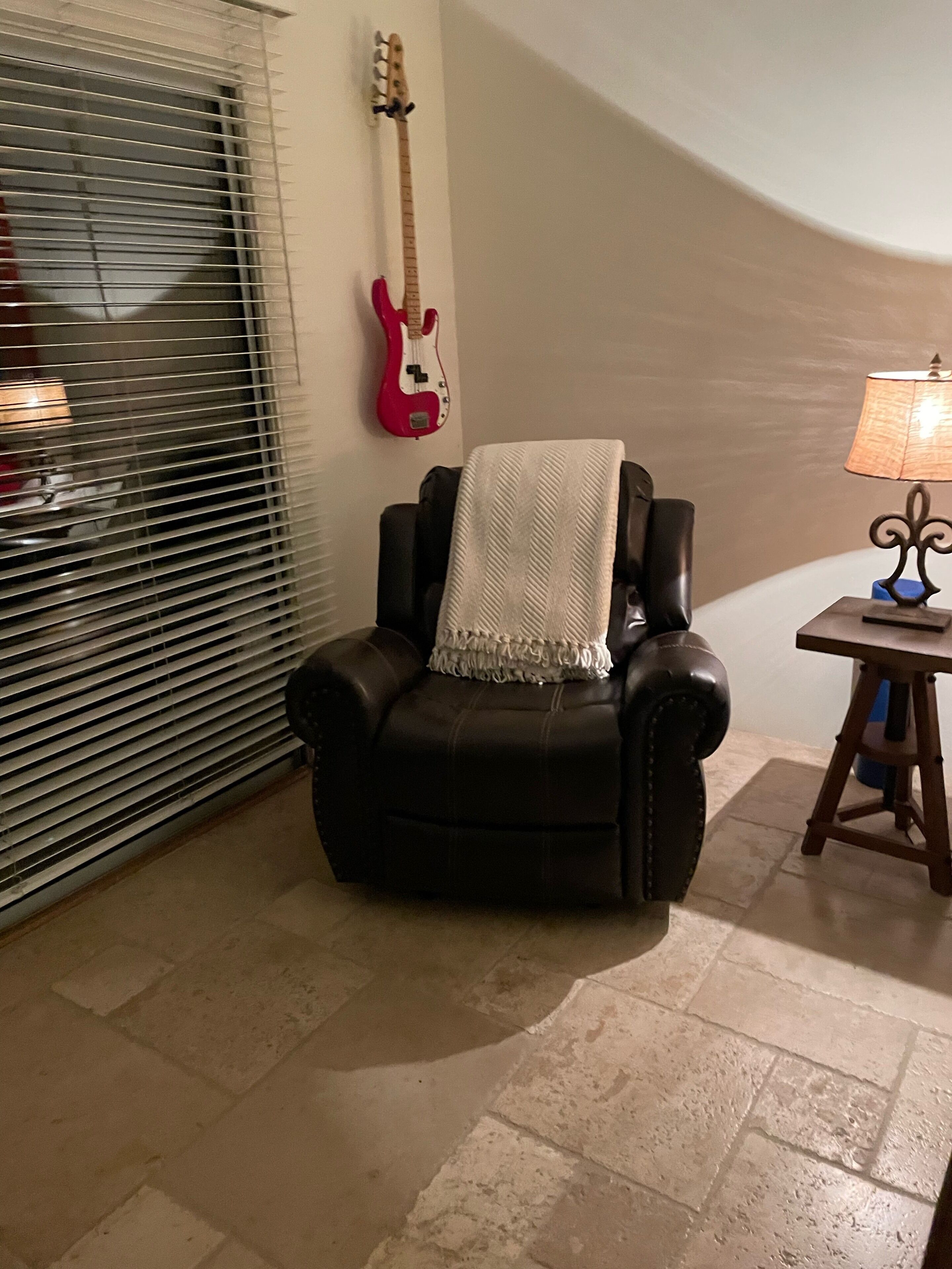 A room with a black leather armchair, a red electric guitar on the wall, a lamp on a wooden table, and a window with blinds.