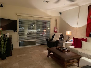 Uma sala de estar com televisão, um violão, um sofá, uma mesa de centro e uma janela com persianas.