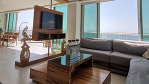 A modern living room with a flat-screen TV, a ceiling fan, and a large window offering a view of the sky. - BEACHFRONT PENTHOUSE with PRIVATE POOL (Nuevo Vallarta)