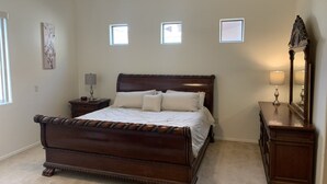A bedroom with a large wooden bed, a nightstand with a lamp, a dresser with a mirror, and three small windows on the wall. - Rancho Vistoso Getaway (Oro Valley)