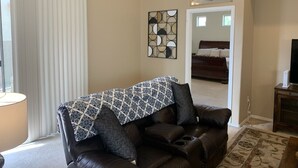 A living room with a brown leather sofa, a patterned throw, and a large window with blinds. - Rancho Vistoso Getaway (Oro Valley)