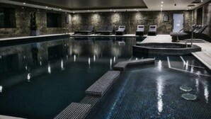 A modern indoor swimming pool with a clear water surface, lounge chairs, and a small hot tub area.