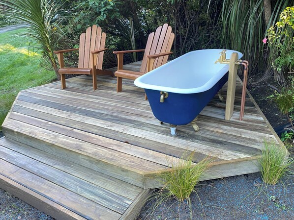 Eine Holzterrasse mit einer blauen Badewanne, zwei Holzstühlen und umgebender Bepflanzung.