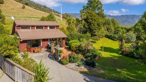 A two-story house with a large porch, surrounded by lush greenery and a view of distant mountains. - Queen Charlotte Secret Oasis, Marlborough Sounds. (Linkwater)