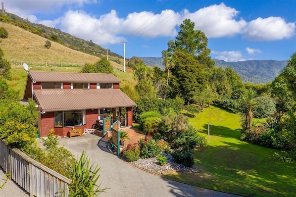 A two-story house with a large porch, surrounded by lush greenery and a view of distant mountains. - Queen Charlotte Secret Oasis, Marlborough Sounds. (Linkwater)