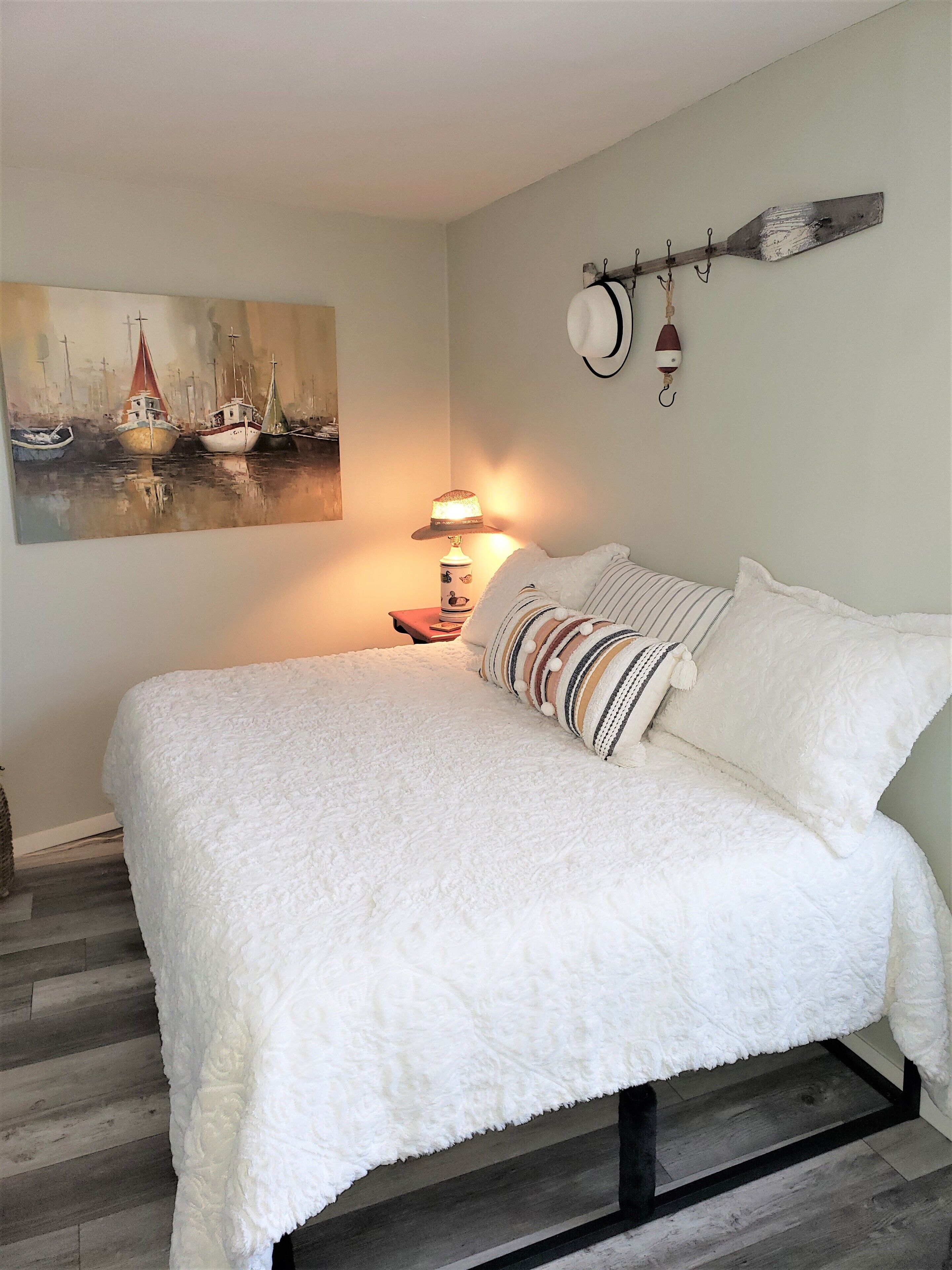 A bedroom with a large bed, a painting of boats on the wall, a lamp, and a wall-mounted hook.