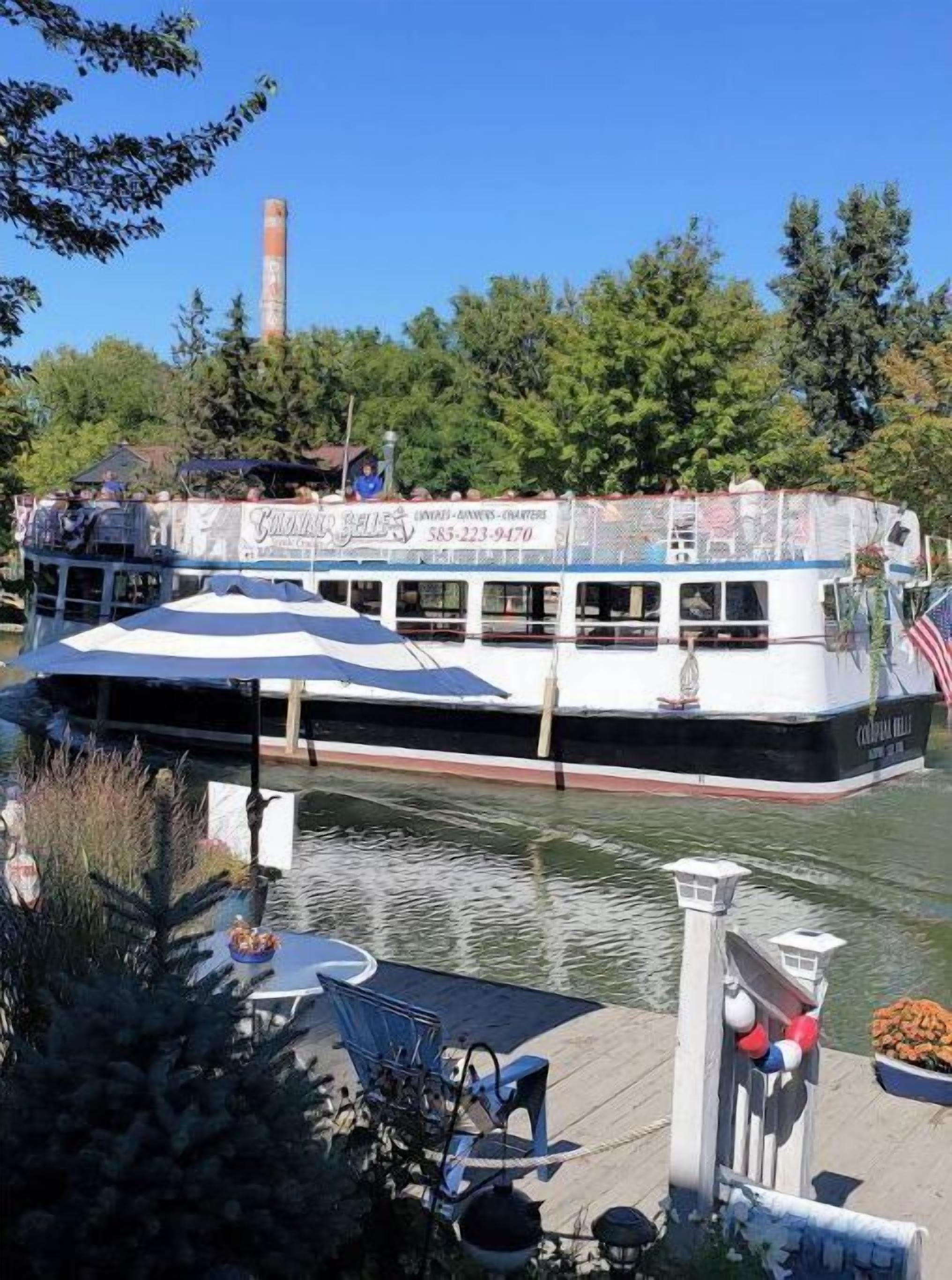 Un bateau fluvial doté d’un pont couvert et arborant le drapeau américain, amarré à un quai agrémenté d’une petite zone d’embarquement et de verdure.